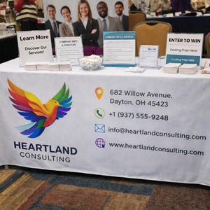 以下が含まれることがあります： カラフルな鳥のデザインが施された、Heartland Consultingのロゴが入った白いテーブルクロス。テーブルには、住所と電話番号を含む情報看板、名刺、連絡先の詳細が表示されています。