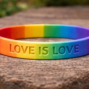 May include: A rainbow-colored silicone bracelet with the words "LOVE IS LOVE" embossed on the front. The bracelet transitions through the colors of the rainbow, from red to purple. The bracelet is resting on a textured, brown surface.