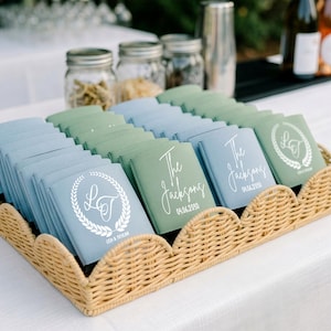 Puede incluir: Una bandeja de mimbre trenzado exhibe recuerdos de boda personalizados. Los pequeños recuerdos cuadrados son de tonos azules y verdes. Cada recuerdo está impreso con un nombre, una fecha y un elemento decorativo blanco. La bandeja está sobre un mantel blanco.