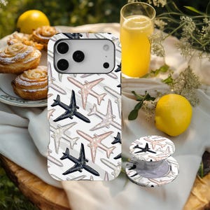 Könnte beinhalten: Eine weiße Handyhülle und ein passender Handyhalter mit einem Muster aus schwarzen, rosa und grauen Flugzeugen. Die Handyhülle liegt auf einer Holzoberfläche mit Gebäck, einem Glas Orangensaft und einer Zitrone.