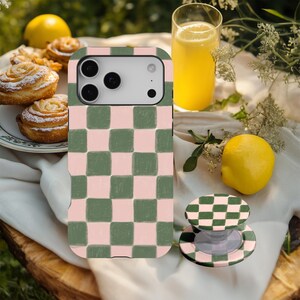 May include: A mobile phone case and phone grip featuring a pink and green chequered design. The phone case is fitted to a phone with multiple camera lenses. The phone grip is round and has a matching pattern. The items are on a wooden surface with food and drinks.