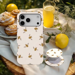 May include: A phone case and pop socket with a repeating pattern of cartoon bees. The case is off-white with brown and yellow bees. A lemon, pastry, and a glass of yellow liquid are also in the image.