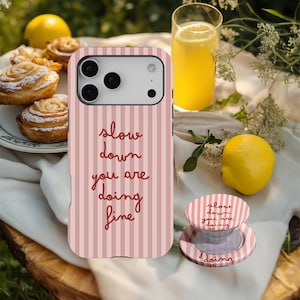 May include: A pink and white striped phone case with the words "slow down you are doing fine" in a cursive font. A matching phone grip is next to the phone case. The image also includes a glass of yellow liquid, lemons, and pastries.