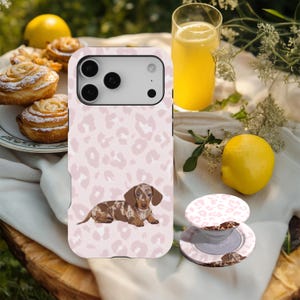 Può includere: Una custodia per telefono rosa con un motivo leopardato e un'illustrazione di bassotto. Un pop-socket abbinato è accanto al telefono. L'immagine include anche un bicchiere di succo d'arancia, un limone e pasticcini.