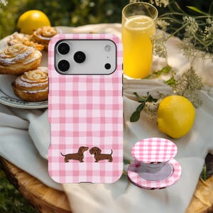 Könnte beinhalten: Rosa karierte Handyhülle mit zwei Dackel-Illustrationen. Ein passender Pop-up-Handyhalter ist ebenfalls zu sehen. Die Hülle und der Halter liegen auf einer weißen Oberfläche mit einer Zitrone, einem Glas Saft und Gebäck im Hintergrund.