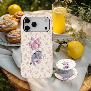 Puede incluir: Funda de teléfono y soporte pop con una ilustración de Eeyore con globos rosas. La funda y el soporte pop tienen un fondo floral crema y rosa, evocando un tema infantil clásico.