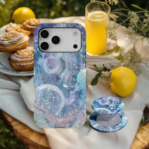 Könnte beinhalten: Smartphone-Hülle und passender Telefonhalter mit einem abstrakten Design in Blau, Lila und Weiß. Die Hülle liegt auf einer weißen Oberfläche mit einer Zitrone, Gebäck und einem Glas Saft.