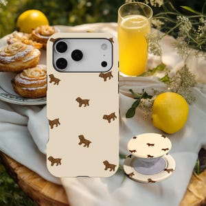 Könnte beinhalten: Eine beige Handyhülle und ein passender Handyhalter mit einem sich wiederholenden Muster aus braunen Hundeillustrationen. Die Handyhülle befindet sich auf einer weißen Oberfläche mit einem Glas gelber Flüssigkeit, einer Zitrone und Gebäck im Hintergrund.