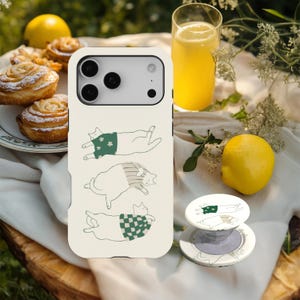 May include: A cream phone case and pop socket with a cat design. The case shows three cats in sweaters, with green and white patterns. A lemon, pastries, and a drink are in the background. The scene is set outdoors.
