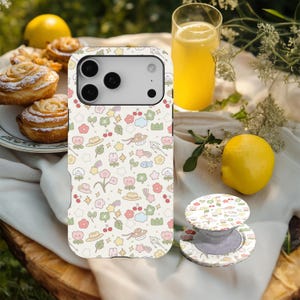May include: A phone case and matching phone grip with a playful design of flowers, hats, cherries, and rainbows in pastel shades. The phone case is on a white surface with a glass of orange juice, a lemon, and pastries in the background.