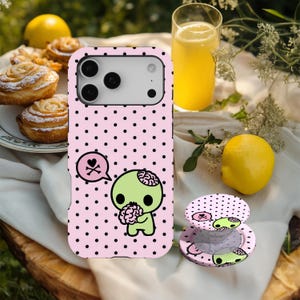 May include: Pink phone case and matching PopSocket with a black polka dot pattern. The case features a green zombie character holding a brain, with a speech bubble and a skull and crossbones. A lemon and pastry are in the background.