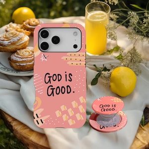 May include: A pink phone case and matching phone grip with the text "GOD IS GOOD." The case and grip have a colourful abstract design with shapes and lines. The image also includes a glass of orange juice, lemons, and pastries.