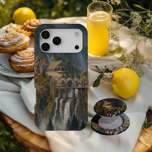 May include: A smartphone case and phone grip featuring a detailed landscape painting of a fantasy architectural structure with waterfalls. The case is dark green with a white camera section. A yellow lemon and pastry are also visible.