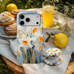 May include: A smartphone case and a phone grip featuring a floral design of white and orange daffodils on a light blue background. The image also includes a glass of orange juice, a lemon, and pastries.