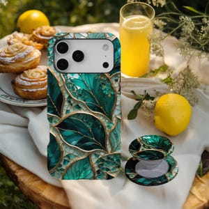 May include: A smartphone case and matching phone grip featuring a mosaic leaf design in shades of teal, gold, and white. The case is on a white surface with a glass of orange juice, lemons, and pastries in the background.