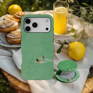 May include: Green phone case with an illustration of two people swimming in water. A matching green pop socket is next to the phone case. The image also includes a glass of orange juice, a lemon, and pastries.