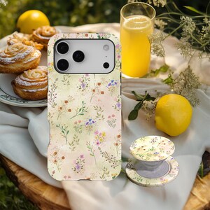 May include: A phone case and phone grip with a floral design, featuring a pastel backdrop and vibrant flowers. The case is on a wooden surface with pastries, a glass of juice, and lemons in the background.