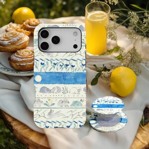 Puede incluir: Una funda de teléfono y un agarre a juego con un diseño caprichoso. La funda presenta patrones florales azules e ilustraciones de conejos sobre un fondo blanco. El agarre del teléfono es redondo y tiene un diseño a juego.