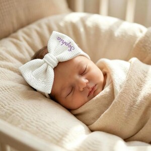 Puede incluir: Un bebé dormido con una diadema blanca con lazo y el nombre "Sophia" bordado en morado. El bebé está envuelto en una suave manta color crema, descansando sobre una superficie color crema.