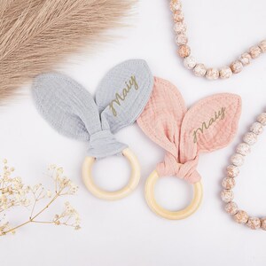 May include: Two baby rattles featuring smooth, round, light-colored wooden rings attached to soft, knotted fabric bunny ears. One set of ears is light gray and the other is dusty pink. Each fabric ear is personalized with the name Maiy in gold.