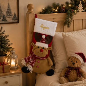 May include: A red Christmas stocking with a white faux fur cuff featuring the embroidered name Ryan in gold thread. The stocking displays a plush tan reindeer character wearing a red and white patterned scarf, a matching hat, and two buttons.