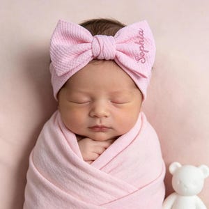 Op de afbeelding: Een lichtroze babyhoofdband met een grote, gestructureerde stoffen strik waarop aan de rechterkant de naam Sophia is geborduurd in een bijpassend roze cursief lettertype. De hoofdband is gemaakt van zacht geribd materiaal en wordt door een baby gedragen.