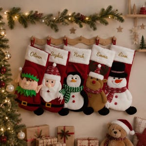 May include: Five red and white Christmas stockings hanging on a wooden rack, each featuring a plush character: an elf, Santa Claus, a penguin, a reindeer, and a snowman. Each stocking has a white cuff embroidered with the names Oliver, Sophia, Noah, Emma, and Liam.