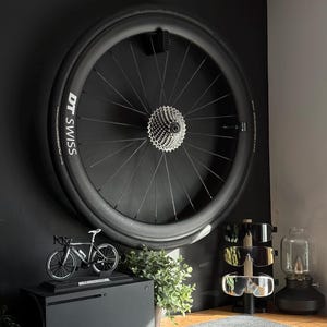 May include: A black bicycle wheel with DT Swiss branding is mounted on a black wall. A miniature bicycle model sits atop a black box, next to a small potted plant. Sunglasses and a lamp are also visible.
