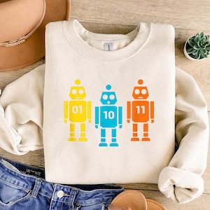 Może przedstawiać: Kremowa bluza z trzema kolorowymi wzorami robotów. Każdy robot jest w innym kolorze: żółtym, niebieskim i pomarańczowym, i każdy ma numer: 01, 10 i 11. Roboty mają prosty, kreskówkowy styl.