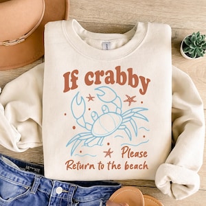 May include: Cream-colored sweatshirt with the text "If crabby Please Return to the beach" and a blue crab illustration. The design includes starfish and dots. The sweatshirt is paired with denim shorts and sandals.