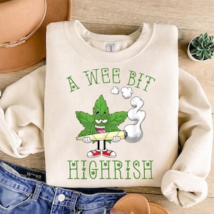 May include: Cream-colored sweatshirt with the text "A Wee Bit Highrish" in green. The graphic features a cartoon cannabis leaf character holding a lit joint, wearing red sneakers, and exhaling smoke.
