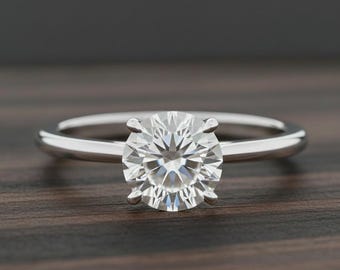 May include: A solitaire engagement ring with a round-cut diamond set in a polished silver band. The diamond is the focal point, held by four prongs, and sparkles against the dark wood background. The ring is a classic design.