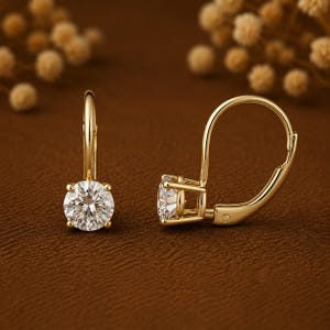 May include: A pair of gold leverback earrings, each featuring a single round-cut clear gemstone held in a four-prong setting. The metal has a polished yellow gold finish, and the design includes a curved wire mechanism for secure closure.