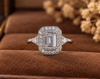 May include: A silver-toned ring featuring a central emerald-cut stone surrounded by a rectangular halo of small round stones. The band includes a triangular stone on each side, connecting to the main setting with a polished metal finish.
