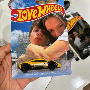 May include: A custom Hot Wheels toy car packaging featuring a photograph of two people embracing. The yellow and black Lamborghini Huracán Sterrato toy car is encased in a clear plastic blister pack. The card displays the text Love Wheels and Lamborghini Huracán Sterrato.
