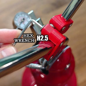 Może przedstawiać: Srebrny metalowy klucz imbusowy z oznaczeniem H2.5 używany przy czerwonym urządzeniu mechanicznym z okrągłym manometrem. Narzędzie jest ustawione tak, aby regulować czerwone plastikowe elementy połączone z błyszczącym chromowanym metalowym prętem.