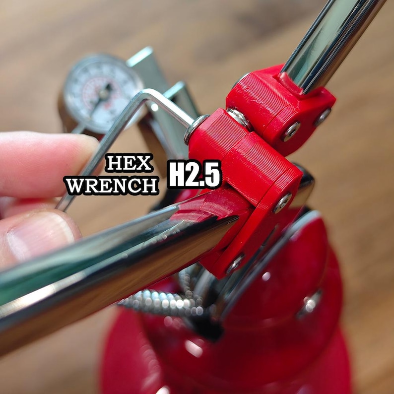 Może przedstawiać: Srebrny metalowy klucz imbusowy z oznaczeniem H2.5 używany przy czerwonym urządzeniu mechanicznym z okrągłym manometrem. Narzędzie jest ustawione tak, aby regulować czerwone plastikowe elementy połączone z błyszczącym chromowanym metalowym prętem.