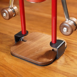 May include: A close-up of a manual espresso maker. The base is made of wood with a black trim. It has two red vertical poles and two wooden handles. The metal arms and handles are silver. The espresso maker is on a wooden surface.