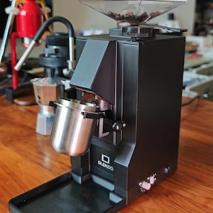 May include: A black electric coffee grinder with a hopper filled with coffee beans. A stainless steel portafilter is positioned to catch the ground coffee. The grinder has the word "SILENZIO" printed on the front.