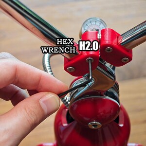 Może przedstawiać: Czarny klucz imbusowy w kształcie litery L jest włożony w czerwony plastikowy zacisk przymocowany do chromowanego metalowego pręta. Tekst HEX WRENCH H2.0 jest widoczny w pobliżu czerwonego zacisku. Urządzenie posiada okrągły wskaźnik i czerwoną podstawę.