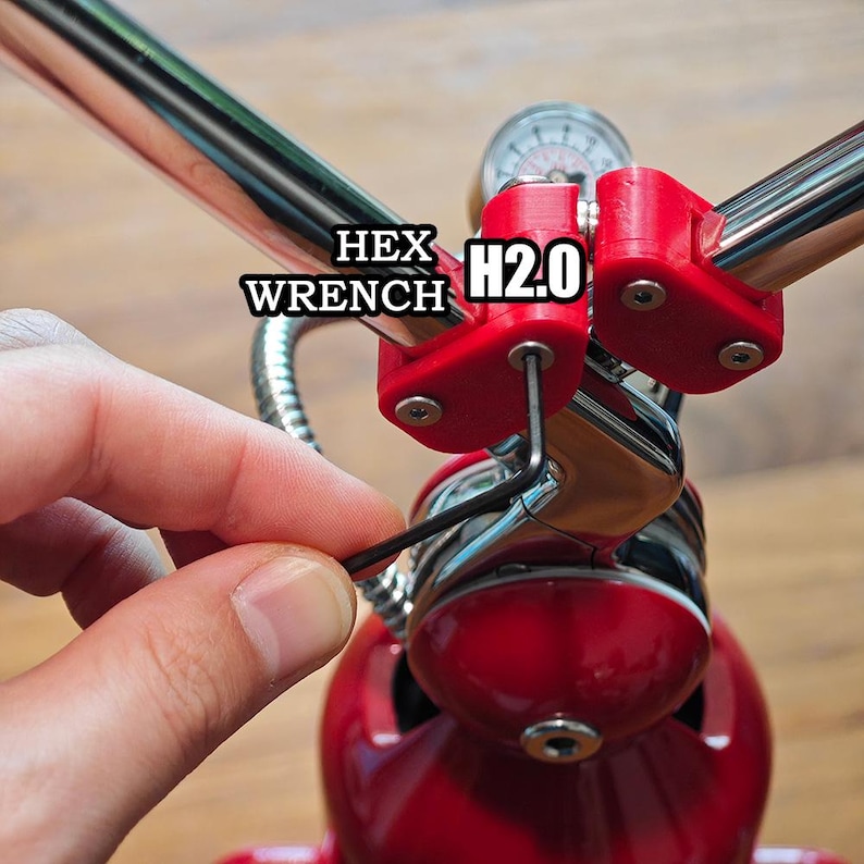 Może przedstawiać: Czarny klucz imbusowy w kształcie litery L jest włożony w czerwony plastikowy zacisk przymocowany do chromowanego metalowego pręta. Tekst HEX WRENCH H2.0 jest widoczny w pobliżu czerwonego zacisku. Urządzenie posiada okrągły wskaźnik i czerwoną podstawę.