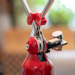 May include: A close-up shot of a red and silver espresso maker. The device has a red base, a silver lever, and a small silver gauge. The design includes a red plastic component and two silver metal rods extending upwards.
