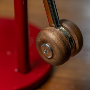 以下が含まれることがあります： 木製のハンドルと銀色の金属アクセントが付いた赤いキッチン家電のクローズアップ。ハンドルは、銀色の金属棒で接続された2つの木製ホイールでできています。家電製品には赤いベースがあります。