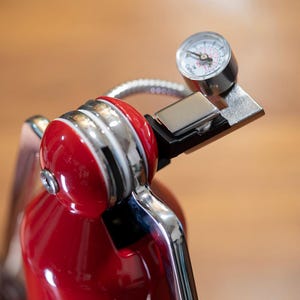May include: Close-up of a red fire extinguisher with chrome detailing. A pressure gauge is visible at the top, showing pressure readings. The extinguisher has a polished finish and a silver hose.
