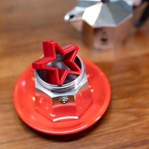 以下が含まれることがあります： 赤い星型のコーヒーメーカーアクセサリーが、赤いコーヒーメーカーのベースの上に置かれています。コーヒーメーカーは銀色で一部が見え、背景には木製の表面があります。星型のアクセサリーはプラスチック製です。
