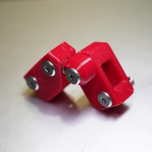 Może przedstawiać: Jaskrawoczerwony, wydrukowany w 3D mechaniczny element zawiasu o teksturowanej powierzchni. Zesp&oacute;ł składa się z dw&oacute;ch połączonych czerwonych plastikowych części, złączonych centralną metalową śrubą oraz dodatkowymi srebrnymi metalowymi wkrętami.