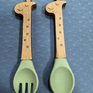 May include: A fork and a spoon set featuring light wood handles carved into the shape of giraffe heads with engraved facial features and spots. The utensil heads are made of smooth, matte light green silicone, providing a soft contrast.