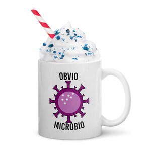 Puede incluir: Taza de cerámica blanca con las palabras "OBVIO MICROBIO" y un diseño de microbio morado. La taza está llena de crema batida y chispas azules, y una pajita a rayas rojas y blancas.
