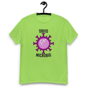 Puede incluir: Una camiseta verde lima con un gráfico de microbio morado y las palabras "OBVIO" y "MICROBIO" en negro. El diseño del microbio presenta un círculo rosa central rodeado de púas moradas.