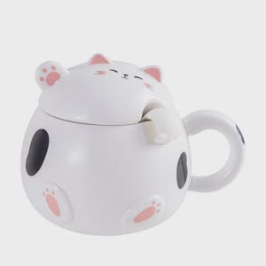 May include: A white ceramic cat-themed mug with a lid. The mug features black spots and pink paw prints. The lid is shaped like a cat's head with pink ears and a paw. The handle has a black spot.